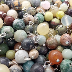 May include: A close-up of a variety of round, smooth, natural stone beads with silver wire loops. The beads are in shades of green, brown, white, and beige.