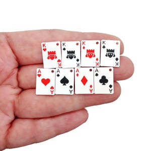 May include: Miniature playing cards featuring the King and Ace of various suits. The cards are white with red, black, and gold details. The cards are held in a hand.