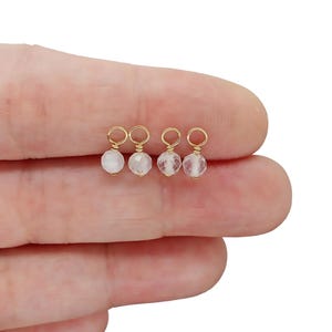 May include: Four small gold-colored charms, each with a small, round, faceted, white bead. The charms are held between fingers, with the gold loops visible. The beads have a translucent quality.