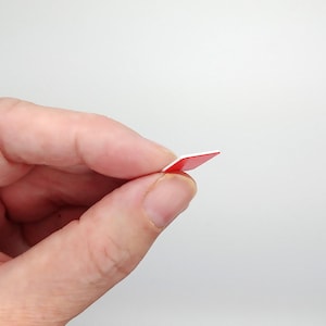 May include: A small, square, red and white sticker held between fingers. The sticker has a bright red top surface and a white edge. The sticker is thin and appears to be made of paper or a similar material. The background is plain white.