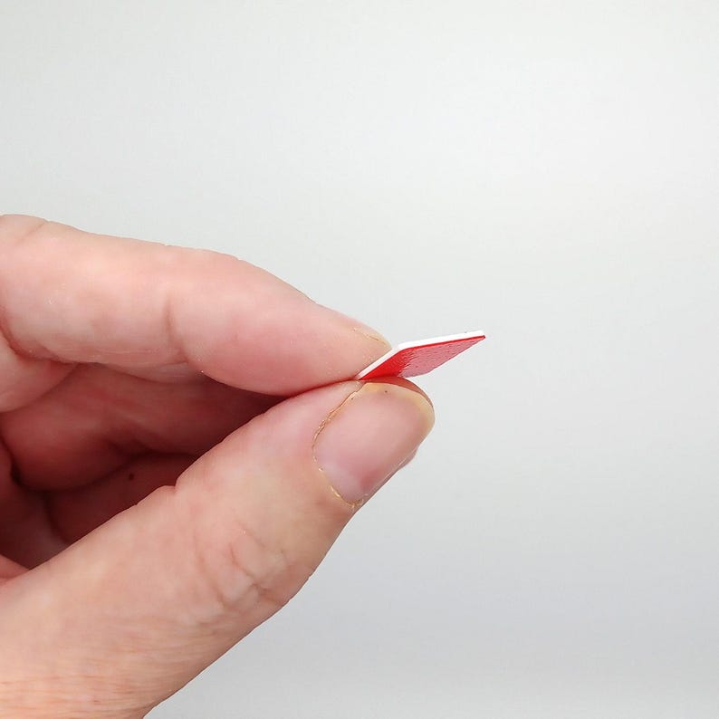 May include: A small, square, red and white sticker held between fingers. The sticker has a bright red top surface and a white edge. The sticker is thin and appears to be made of paper or a similar material. The background is plain white.