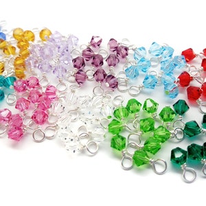 May include: A collection of colorful crystal beads in various shades, including blue, yellow, purple, pink, and green. Each bead is attached to a silver wire loop, suggesting they are components for jewelry making or other crafts. The beads are arranged on a white background.