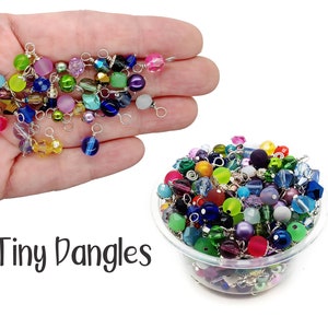 Peut inclure: Un bol rempli de perles de verre colorées avec des anneaux de saut argentés. Les perles sont de formes et de tailles variées. Le texte "Tiny Dangles" est écrit sous le bol.