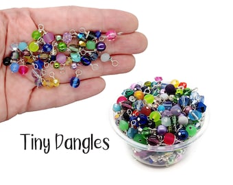 May include: A bowl filled with colourful glass beads with silver jump rings. The beads are various shapes and sizes. The text "Tiny Dangles" is written below the bowl.