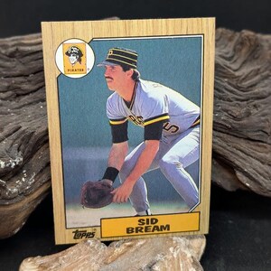 Puede incluir: Una tarjeta de béisbol Topps vintage con Sid Bream de los Piratas de Pittsburgh. La tarjeta tiene un diseño de marco de madera y muestra a Bream con su uniforme de los Piratas, con el logotipo del equipo en la esquina superior izquierda. La tarjeta está en buen estado.