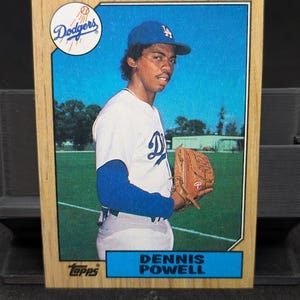 May include: A vintage baseball card featuring Dennis Powell of the Los Angeles Dodgers. The card has a wooden frame and a blue and white color scheme. The Dodgers logo is visible, along with the Topps logo and Powell's name.