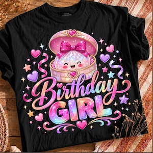 May include: A black t-shirt featuring a graphic of a smiling, glittery character inside an open, ornate pink jewelry box. The text "Birthday GIRL" appears below in a colorful, gradient font, surrounded by pink hearts, stars, and ribbons.