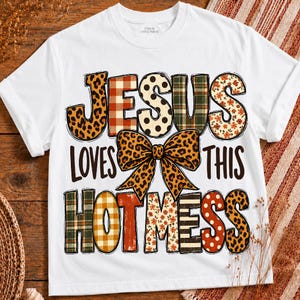Peut inclure: Un t-shirt blanc à manches courtes avec le texte 'JESUS LOVES THIS HOT MESS' en grandes lettres à motifs variés. Le design comprend un nœud central à imprimé léopard et divers motifs comme des carreaux, des pois et des fleurs dans les lettres.