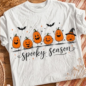Peut inclure: Un t-shirt blanc avec un motif de six citrouilles orange aux expressions et postures variées. Les citrouilles sont accompagnées de chauves-souris noires, de petites étoiles orange et du texte cursif noir "spooky season" en dessous.
