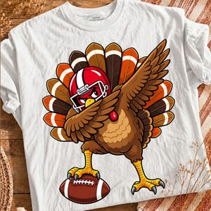 Könnte beinhalten: Ein weißes T-Shirt mit einer Grafik eines braunen Truthahns, der einen roten Football-Helm trägt und dabbt. Der Truthahn hat einen großen, bunten Schwanz mit braunen, orangefarbenen und weißen Federn. Eine Kralle ruht auf einem braunen Football.
