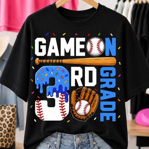 May include: A black t-shirt featuring the text GAME ON 3RD GRADE in white and blue. The design includes a baseball, a wooden baseball bat, a large number three with blue icing and sprinkles, and a brown leather baseball glove with a baseball.