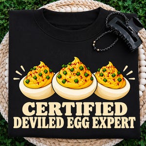 Peut inclure: Un sweat-shirt noir à col rond orné d'un motif représentant trois œufs farcis garnis de ciboulette verte et de paprika rouge. Sous les œufs, le texte "CERTIFIED DEVILED EGG EXPERT" est inscrit en grandes lettres capitales claires.