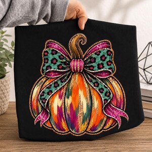 Peut inclure: Un t-shirt noir avec un motif de citrouille à la surface texturée orange, jaune et violette. La citrouille est ornée d'un grand nœud sarcelle à motif léopard noir avec des bords roses scintillants, surmonté d'une tige de couleur marron.