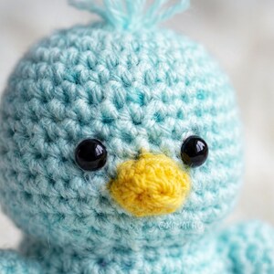 Puede incluir: Primer plano de un p&aacute;jaro de ganchillo hecho a mano con cuerpo azul claro, ojos negros y pico amarillo. El p&aacute;jaro tiene un aspecto texturizado y cosido y un peque&ntilde;o mech&oacute;n de hilo azul en la cabeza. Un art&iacute;culo artesanal hecho a mano.