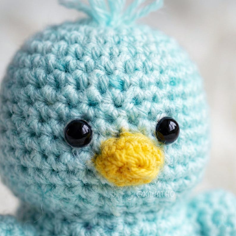Puede incluir: Primer plano de un p&aacute;jaro de ganchillo hecho a mano con cuerpo azul claro, ojos negros y pico amarillo. El p&aacute;jaro tiene un aspecto texturizado y cosido y un peque&ntilde;o mech&oacute;n de hilo azul en la cabeza. Un art&iacute;culo artesanal hecho a mano.