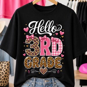 May include: A black short-sleeve t-shirt featuring the white text Hello and the pink and leopard print text 3RD GRADE. The design includes pink bows on the numbers, small pink hearts, and gold stars, all centered on the front of the garment.