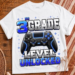 May include: A white t-shirt featuring a graphic print of a black video game controller centered between the text 3RD GRADE and LEVEL UNLOCKED. The text is styled in a metallic blue and silver font, surrounded by small blue stars and pixelated accents.