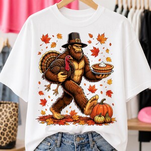 Peut inclure: Un t-shirt blanc avec un motif représentant un Bigfoot brun portant un chapeau de pèlerin, tenant une dinde sous un bras et une tarte à la citrouille dans l'autre main. Le design inclut des feuilles d'automne orange et rouges et deux citrouilles.