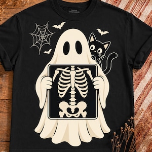 May include: A black t-shirt featuring a graphic of a cream-colored ghost holding an X-ray of a human rib cage and pelvis. A small black cat stands beside the ghost, with a spider web and three small bats positioned in the upper background area.