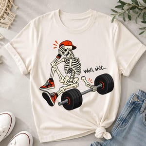 May include: A cream-colored t-shirt featuring a graphic of a skeleton wearing a red baseball cap and orange and black sneakers. The skeleton sits next to a barbell with one detached arm, and the text "Well, shit..." is printed in black.