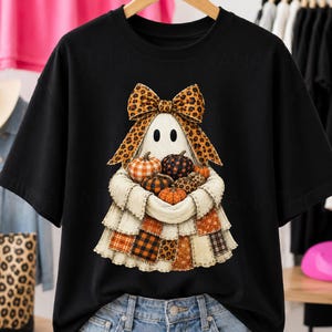 Könnte beinhalten: Ein schwarzes Kurzarm-T-Shirt mit einer Grafik eines weißen Geistes, der eine große Schleife mit Leopardenmuster trägt. Der Geist hält eine Ansammlung kleiner Kürbisse mit verschiedenen Mustern, darunter orangefarbenes Karo und schwarze Punkte.