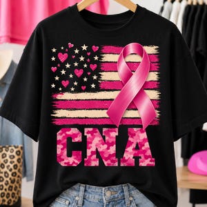 Peut inclure: Un t-shirt noir avec un motif de drapeau américain stylisé aux rayures roses et crème, agrémenté de cœurs roses et d'étoiles blanches. Un grand ruban de sensibilisation rose est superposé. Les lettres CNA sont en motif camouflage rose.