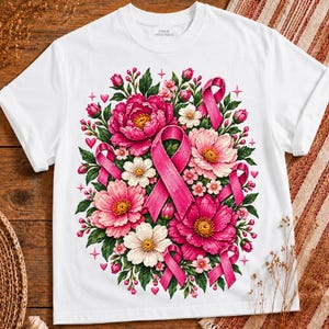 May include: A white t-shirt featuring a central graphic of vibrant pink and white flowers interspersed with multiple pink awareness ribbons. The design includes small pink hearts and star accents, creating a detailed floral arrangement with green leaves.