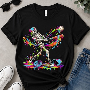 May include: A black short-sleeved t-shirt featuring a graphic of a skeleton wearing a baseball cap and trainers, swinging a bat at a baseball. The design includes vibrant, multi-coloured paint splatters in neon pink, blue, green, and orange.