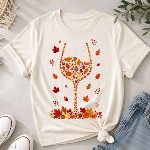 Peut inclure: Un t-shirt à manches courtes de couleur crème arborant un motif en forme de verre à vin composé d'éléments automnaux variés, notamment des feuilles orange, rouges et jaunes, des petites citrouilles, des glands et des pommes de pin.