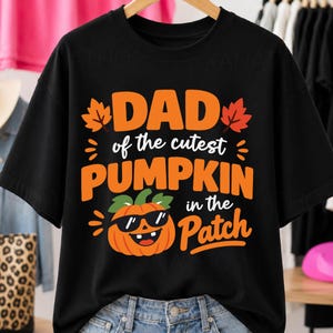 Peut inclure: Un t-shirt noir à manches courtes avec un texte orange et blanc indiquant DAD of the cutest PUMPKIN in the Patch. Le motif comprend un dessin de citrouille orange portant des lunettes de soleil noires, entouré de deux feuilles d'érable orange.