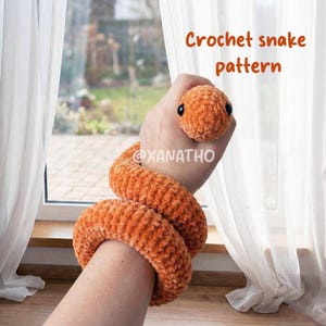 Peut inclure: Un serpent orange crocheté avec des yeux noirs est enroulé autour d'une main. Le corps du serpent est enroulé et la tête est positionnée près des doigts. Le texte "Crochet snake pattern" est visible dans le coin supérieur droit.