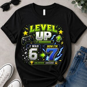 Könnte beinhalten: Ein schwarzes Kurzarm-T-Shirt mit einer bunten Grafik und dem Text LEVEL UP, I WAS 6, NOW I'M 7, UNLOCKED: AWESOME. Das Design enthält einen Videospiel-Controller, einen Fortschrittsbalken, Blitze, Sterne und ein Trophäen-Symbol.
