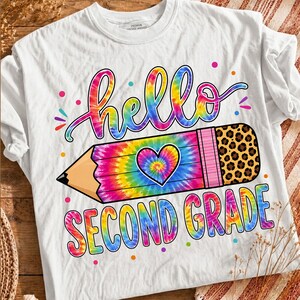 Peut inclure: Un t-shirt blanc avec un motif tie-dye coloré représentant un crayon avec un cœur au centre. Le texte hello est écrit en police cursive tie-dye au-dessus du crayon et SECOND GRADE en police bloc tie-dye en dessous, avec des détails léopard.
