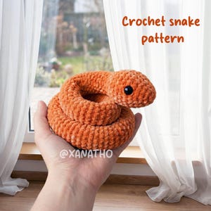 Peut inclure: Un serpent en crochet orange avec un œil noir, enroulé dans une main. Le serpent est fait d'un fil doux et texturé. Le texte de l'image indique "Crochet snake pattern".