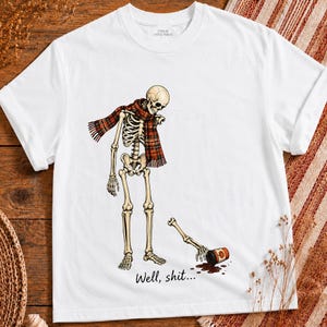 Könnte beinhalten: Ein weißes Kurzarm-T-Shirt mit einer Grafik eines Skeletts, das einen rot-schwarzen Schal trägt. Ein Arm des Skeletts liegt abgetrennt auf dem Boden neben einer umgekippten Dose mit Kürbis-Etikett. Der Text lautet: Well, shit...