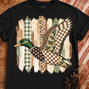 Peut inclure: Un t-shirt noir à manches courtes avec un motif de canard colvert en vol. Le corps du canard est composé de divers motifs, dont des pois marron, un damier marron et blanc, un imprimé léopard et du camouflage. Le fond présente un design de coups de pinceau avec des rayures vichy vertes et beige.