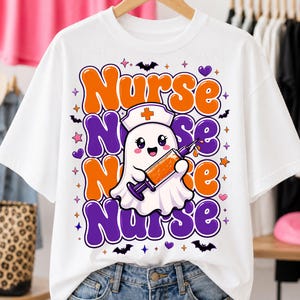 May include: A white t-shirt featuring a graphic of a cute white ghost wearing a nurse cap and holding a syringe filled with orange liquid. The background text reads Nurse in repeating orange and purple wavy bubble letters with small bats and stars.