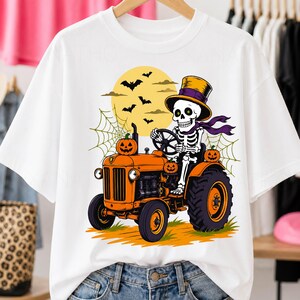 Peut inclure: Un t-shirt blanc avec un motif représentant un squelette portant un haut-de-forme et une écharpe violette, conduisant un tracteur orange. Le dessin inclut une pleine lune jaune, des chauves-souris noires, des toiles d'araignée et trois citrouilles.