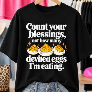 Peut inclure: Un t-shirt noir avec le texte blanc Count your blessings, not how many deviled eggs I'm eating. Le motif comprend un graphique de trois moitiés d'œufs farcis avec des jaunes jaunes et des éclats de paprika orange.