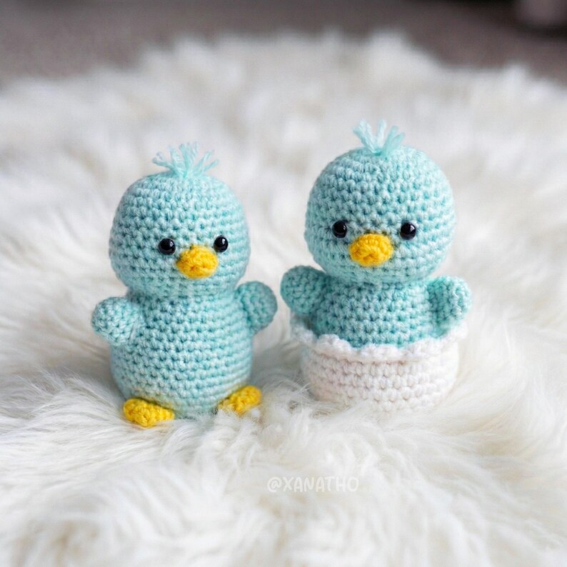 Puede incluir: Dos p&aacute;jaros azules de ganchillo hechos a mano con picos y patas amarillas. Un p&aacute;jaro est&aacute; de pie, el otro est&aacute; en una taza de ganchillo blanca. Los p&aacute;jaros tienen ojos negros y peque&ntilde;os mechones de hilo azul en la cabeza. Sobre una superficie blanca y esponjosa.