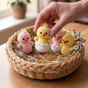 Puede incluir: Una cesta de mimbre contiene varios pollitos de ganchillo en tonos amarillos y rosas, con picos naranjas y ojos negros. Un pollito sale de una c&aacute;scara de huevo blanca. La cesta est&aacute; forrada con musgo y trigo, y una mano alcanza a uno de los pollitos.