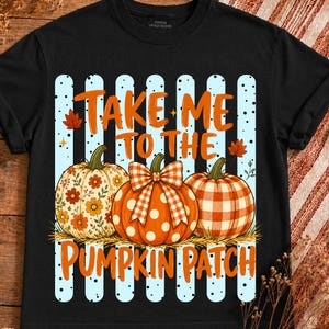 Peut inclure: Un t-shirt noir avec un motif de trois citrouilles posées sur du foin. Les citrouilles présentent des motifs floraux, à pois et à carreaux. Le texte "TAKE ME TO THE PUMPKIN PATCH" est écrit en orange sur des rayures verticales bleu clair.