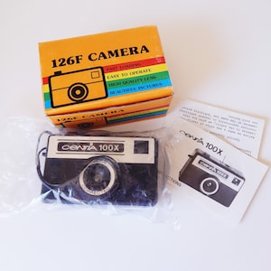 May include: A black and silver 126F camera with a 'CENTIA 100X' label. The camera is in its original packaging, which is orange and yellow with the text '126F CAMERA' and 'BEAUTIFUL PICTURES'.