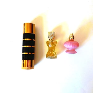 May include: Three vintage perfume bottles. A tall, gold-colored bottle with black bands, and two smaller bottles: one yellow, hourglass-shaped with a silver cap, and a pink, bulbous bottle with a gold-colored top.
