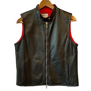 May include: Black leather vest with a red interior lining. The vest has a stand-up collar, a full-length zipper, and two front pockets. The vest is hanging on a wooden hanger.