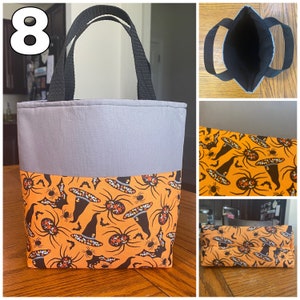 May include: A gray and orange Halloween-themed tote bag with a spider and witch hat pattern. The bag has two black handles and a gray fabric top.