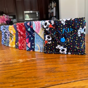 May include: A set of nine small fabric pouches with different colorful patterns. The pouches are lined up in a row on a wooden surface. Each pouch has a flap that folds over and closes with a button.