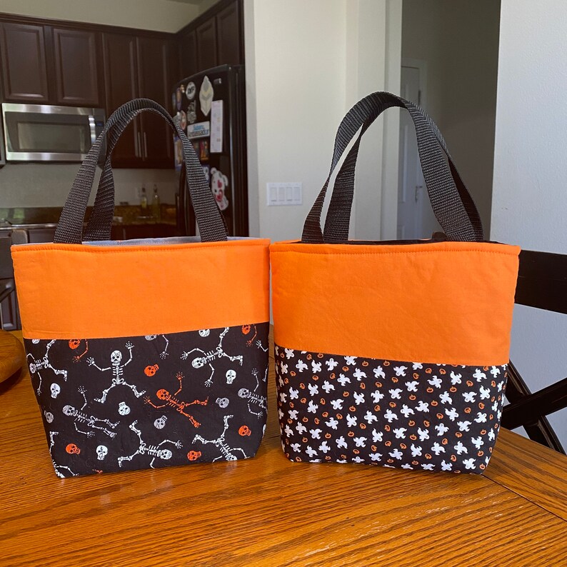 May include: Two Halloween trick-or-treat bags. One bag is orange with a black and white skeleton pattern. The other bag is orange with a black and white ghost pattern.