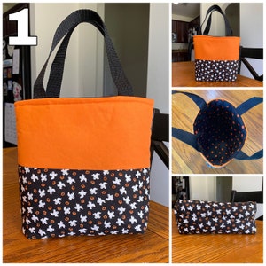 May include: A small orange and black Halloween trick-or-treat bag with a black handle. The bag is made of fabric and has a pattern of ghosts and pumpkins on the bottom half. The bag is sitting on a wooden table.