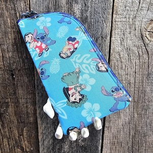 Może przedstawiać: Niebieska kosmetyczka zapinana na zamek błyskawiczny z powtarzającym się wzorem postaci Stitch i Lilo z filmu Disneya Lilo & Stitch. Postacie są ubrane w hawajskie stroje i otoczone są ananasami i kwiatami.